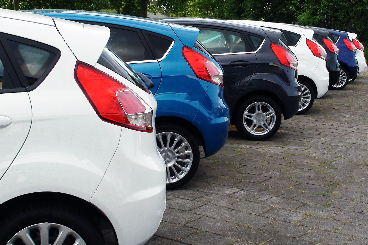 A row of parked cars in various colors, including white, blue, and black, lined up on a paved area with trees in the background.