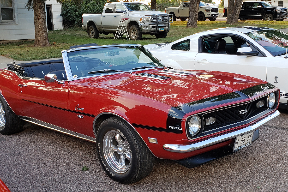 Red vintage convertible sports car parked on street next to a white car and a parked truck in the background.