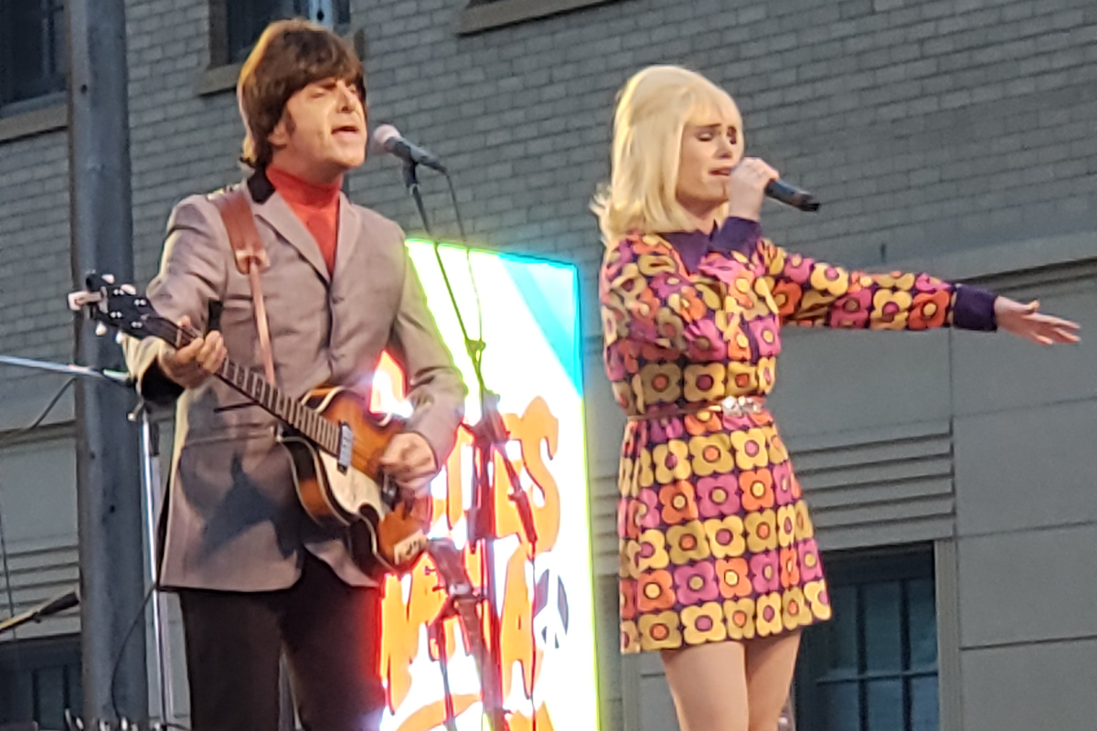 Two performers on stage, one with a guitar dressed in a gray suit, and the other singing, wearing a colorful, floral-patterned dress. A brightly lit sign is visible in the background.