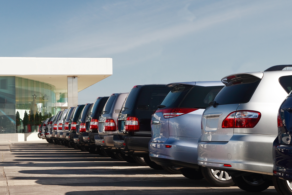 A row of parked cars in a dealership lot, lined up evenly under a clear blue sky with a modern glass building in the background.