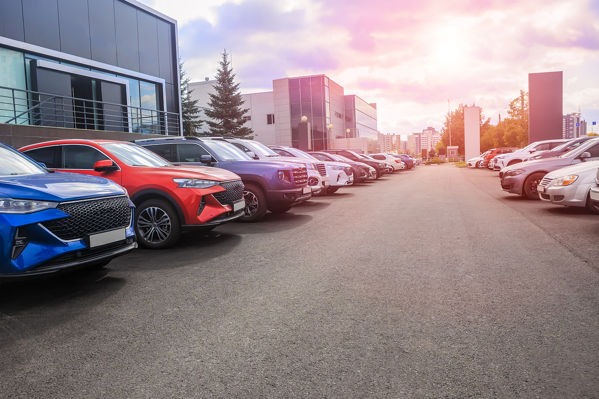 A row of parked cars lines both sides of an asphalt lot, with modern buildings and a partly cloudy sky in the background.