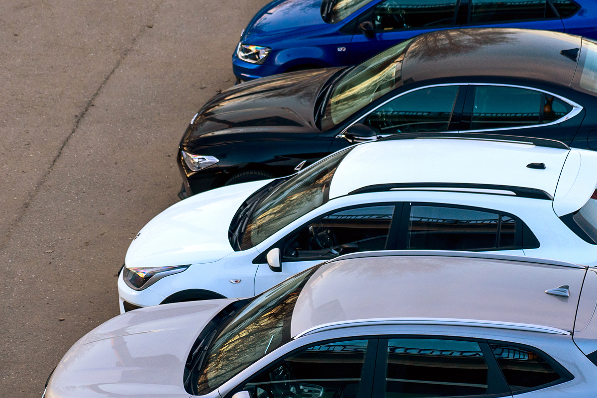 Five parked cars in various colors, including blue, black, white, and beige, are aligned closely in a parking area on asphalt.